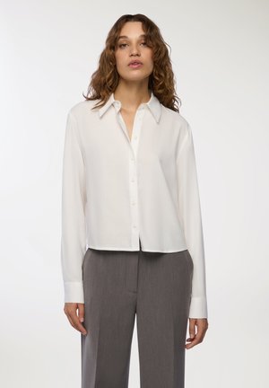 Camicetta bianca a maniche lunghe con bottoni, realizzata in un tessuto morbido; presenta un colletto classico e un orlo cropped, abbinata a pantaloni grigi su misura.