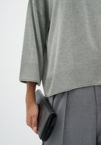 Light grey oversized sweater with ribbed cuffs, paired with gray pleated trousers. A black leather clutch is held in the right hand.