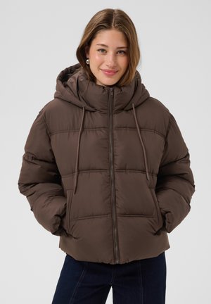 Jeune femme souriante, portant une veste matelassée à capuche marron, les mains dans les poches, debout devant un fond clair uni.