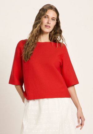 Woman with long wavy hair wearing a bright red loose top with elbow-length sleeves and a white skirt, standing against a plain background.