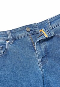 Blå denimjeans med delvis öppen dragkedja och knäppning, som visar gult tyg inuti midjebandet.