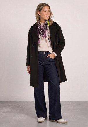 Une femme souriante présente en studio un manteau noir, un pull crème, une écharpe à motifs, un jean large et des baskets blanches.