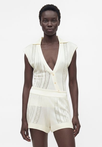 Γυναίκα που φοράει ένα κρεμ πλεκτό αμάνικο τοπ με βαθύ V-λαιμόκοψη και αντιστοιχισμένα σορτς με ψηλή μέση και υφασμένα κάθετα μοτίβα.