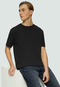 T-shirt noir à manches courtes avec un col rond, fabriqué en tissu doux. Porté avec un jean en denim bleu. Le modèle est adossé à un bloc blanc.