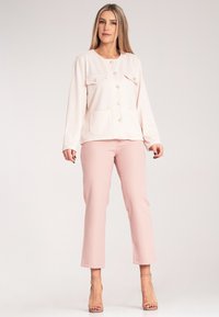 Jasnoróżowa kurtka o teksturze z dwiema kieszeniami, zapięciem na guzik i długimi rękawami, w zestawieniu z dopasowanymi jasnoróżowymi spodniami i sandałkami w kolorze nude.