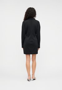 Μαύρο φόρεμ α μακρυμάνικο με γιακά, με λεία υφή και εφαρμοστή σιλουέτα, που φτάνει πάνω από το γόνατο. Ορατά τα τακούνια.