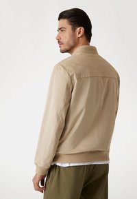 Beige bomberjack met een gladde textuur, geribbelde kraag en manchetten, en naaddetails, gedragen over een wit shirt en groene broek.