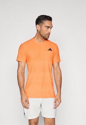 Oranje sportshirt met een gestructureerd patroon, korte mouwen en een zwart logo op de linkerborst. Gecombineerd met witte korte broek.