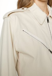 Béžová užitková bunda se strukturovaným límcem, zipovou kapsou na prsou a ramenními epauletkami. Obsahuje růžové prošívání podél švů.