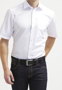 Valkoinen lyhythihainen nappipaita, jossa on rintatasku, yhdistettynä tummansinisiin farkkuihin ja mustaan vyöhön, jossa on hopeinen solki.