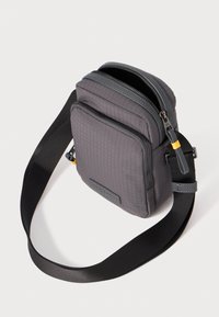 Bolsa cruzada de nylon gris con un patrón de cuadrícula texturizado, dos compartimentos con cremallera y una correa negra ajustable. Acentuada con herrajes amarillos.