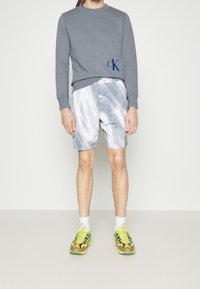 Sweatshirt gris avec logo bleu, associé à un short gris clair tie-dye. Porté avec des chaussettes blanches et des baskets jaunes vives.