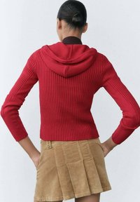 Pull rouge côtelé avec capuche, coupe ajustée, manches longues, associé à une jupe en velours côtelé beige avec des plis et un détail de bouton.