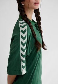 Ung person med langt flettehår som har på seg en grønn kortermet skjorte med hvite sikksakkstriper på ermet.