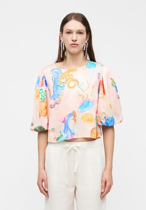 Žena v pastelovej ružovej blúzke s farebnými potlačami morských tvorov, s nafúknutými krátkymi rukávmi a bielymi nohavicami so sťahovacou šnúrkou, na jednoduchom pozadí.