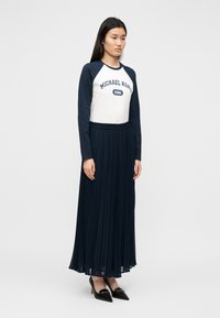 Μακρύ πλισέ φούστα σε ναυτικό μπλε, συνδυασμένη με λευκό και ναυτικό μπλε μακρυμάνικο τοπ με το λογότυπο "Michael Kors 1981" στο κέντρο. Μαύρα τακούνια ολοκληρώνουν το σύνολο.