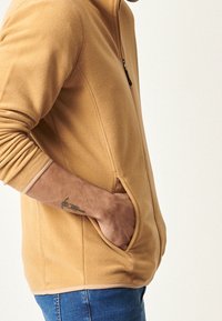 Giacca in pile di un caldo colore sabbia con chiusura a zip, tasche laterali e dettagli a contrasto. Caratterizzata da una vestibilità comoda e rilassata e una texture morbida.