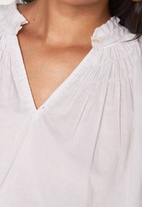 Blusa bianca con scollatura a V, caratterizzata da dettagli arricciati attorno al collo e delicata trina. Tessuto morbido e leggero.