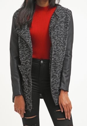 Žena v šedém a černém texturovaném kabátu s černými koženými rukávy, červeném žebrovaném svetru a černých roztrhaných džínách.