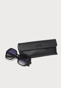 Ochelari de soare hexagonali negri cu lentile degradate, însoțiți de un toc texturat din piele ecologică neagră cu un logo „GUESS” embrosat.