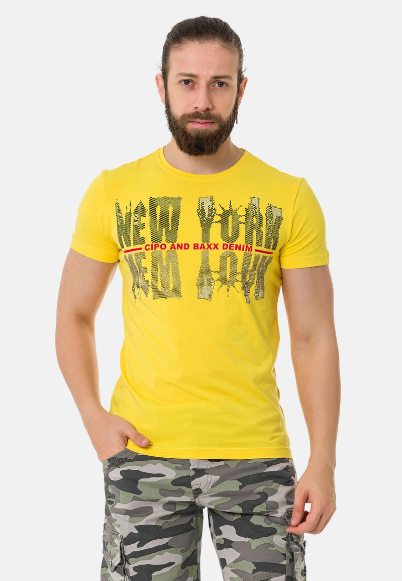 Gelbes kurzärmeliges T-Shirt mit schwarzem und rotem Grafiktext "NEW YORK" und Markennamen, kombiniert mit grauen Camo Cargo-Shorts.