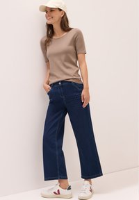 Béžový top s krátkým rukávem v kombinaci s tmavomodrým širokým džínovým kalhotami a bílými teniskami s akcenty. Modelka stojí na světlejším pozadí.