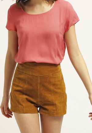 Femme portant un chemisier rose à manches courtes rentré dans un short en suède marron taille haute, debout avec les bras légèrement écartés.