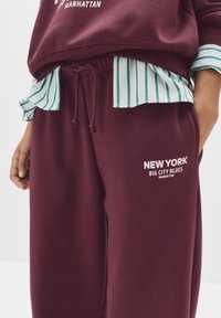 Bordové teplákové nohavice s šnúrkou v páse, s bielym grafickým textom "NEW YORK BIG CITY BLUES MANHATTAN." Prehodené s tričkom so zeleno-bielymi pruhmi.