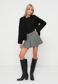 Cardigan noir en tricot avec des accents métalliques, haut noir ajusté, jupe plissée grise et bottes noires à talons hauts. Textures lisses et design structuré.