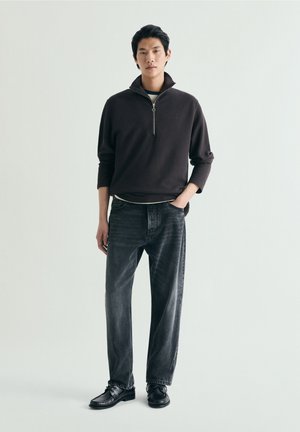 Joven de pie en una postura relajada, vistiendo un jersey negro con cremallera hasta la mitad, vaqueros lavados oscuros y zapatos de cuero negros sobre un fondo liso.