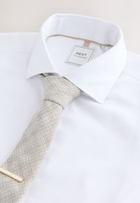 Camicia bianca con colletto classico e cravatta beige chiaro con trama. Presenta una piccola molletta per cravatta dorata e un'etichetta con marchio sul colletto.