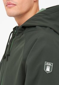 Dunkelgrüne Kapuzenjacke aus glattem Stoff, mit einem markierten Patch auf der Schulter und verstellbaren Kordelzügen am Halsbereich.