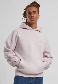 Haut à capuche rose clair en matériau doux, avec une poche kangourou, des poignets côtelés et un logo brodé sur la poitrine.