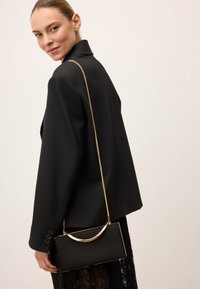 Čierna kabelka na ruku so zlatými detailami, obdĺžnikového tvaru a zakrivenou rúčkou. Textúra látky kontrastuje s pevnou čiernou sakom.
