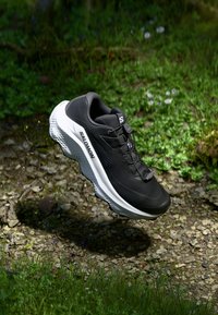 Chaussure de sport noire avec une partie supérieure texturée, une semelle intermédiaire blanche et un design de semelle distinctif. Elle dispose d'un système de laçage et d'une marque sur le côté.