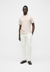 Homme noir debout portant un polo beige à manches courtes, un pantalon blanc et des chaussures habillées noires sur un fond blanc uni.