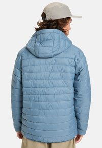 Ljusblå pufferjacka med quiltat mönster, huva och ribbade manschetter. Materialet verkar slätt och isolerat.
