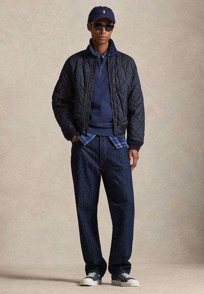 Mand iført navy quiltet jakke, blå sweatshirt med halv lynlås, ternet skjorte, mørke jeans, sneakers, navy kasket og solbriller foran en beige baggrund.