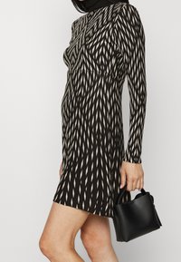 Femme portant une robe longue à manches longues noire et beige à motifs, tenant un petit sac à main noir devant un fond uni.