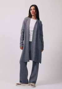 Femme debout portant un cardigan gris ouvert devant, un pantalon large assorti, une chemise blanche et des baskets blanches et marron sur un fond uni.