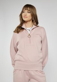 Jasnoróżowy sweter z zamkiem błyskawicznym w stójce, prążkowanymi mankietami i luźnym krojem. Posiada złoty pierścień przy zamku.