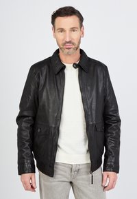 Zwart leren jack met een kraag, ritssluiting aan de voorkant, twee zakken en knoopaccenten. Draagt over een lichtgekleurd gestructureerd vest.