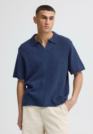 Junger Mann mit kurzem dunklem Haar, der ein marineblaues, strukturiertes Strick-Kurzarm-Poloshirt und hellbeige Hosen trägt.