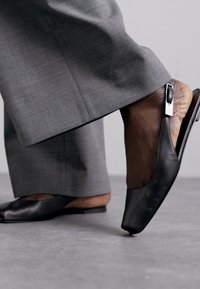 Svart slingback-sko med kvadratisk tå, slät lädermaterial och metallisk detalj på klacken. Bärs med grå skräddarsydda byxor.