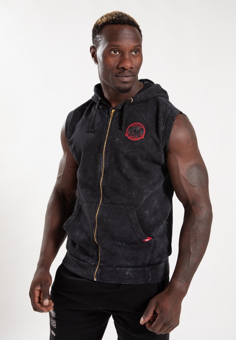 Zwarte mouwloze hoodie met een ritssluiting aan de voorkant, textuurstof, voorzakken en een rode ronde logo op de borst. Gouden rits hardware.