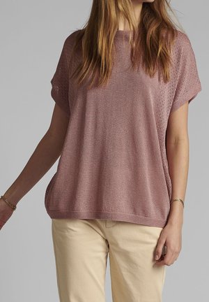 Różowy, krótki sweterek z krótkim rękawem o luźnym kroju, fakturowanym wzorze i szerokim dekolcie. W zestawie z jasnobeżowymi spodniami.