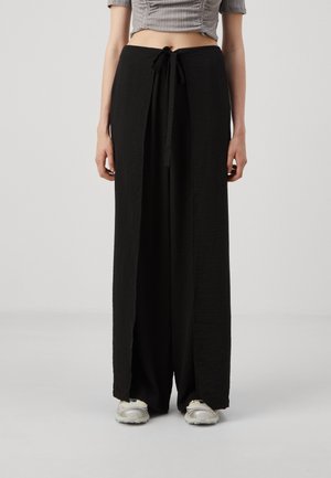 Black wide-leg pants made of textured fabric, featuring a drawstring waist and side slits. Paired with light-colored athletic shoes.