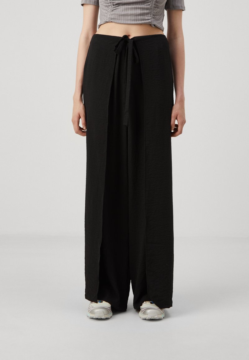 Black wide-leg pants made of textured fabric, featuring a drawstring waist and side slits. Paired with light-colored athletic shoes.