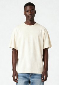 Bēšas kokvilnas t-krekls ar brīvu piegriezumu, apaļu kaklu un īsām piedurknēm. Valkāts kopā ar gaiši ziliem džinsiem. Minimālistisks dizains, maiga tekstūra.