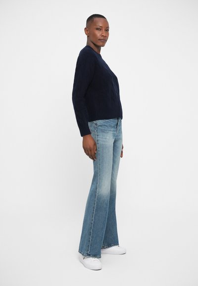 Navy knitted sweater with a textured cable pattern, paired with light blue flared jeans and white sneakers, shown in side profile.